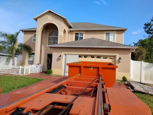 Champs dumpster coming off the trailer onto the wide driveway apron of a two story Clermont home with a white picket fence alongside