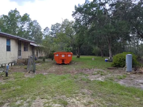 Champs dumpster set on the back lawn of an older Poinciana home during light rain with pines and oaks behind the property