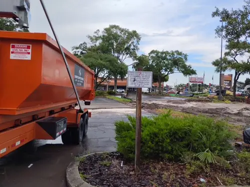 Champs truck with a loaded dumpster parked in a Poinciana retail plaza lot beside a torn out section of asphalt and bare sand
