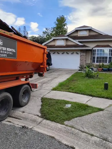 Champs truck and loaded dumpster at the apron of a Mims driveway with the homeowner van parked in front of the tan stucco home
