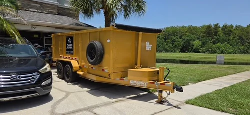 Yellow Champs dump trailer parked on a Paisley driveway alongside the homeowner SUV with the garage open and a pond behind the house