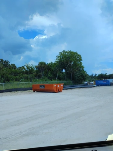 Orange Champs dumpster on the fenced dirt construction yard in Winter Park with stacked pipe nearby