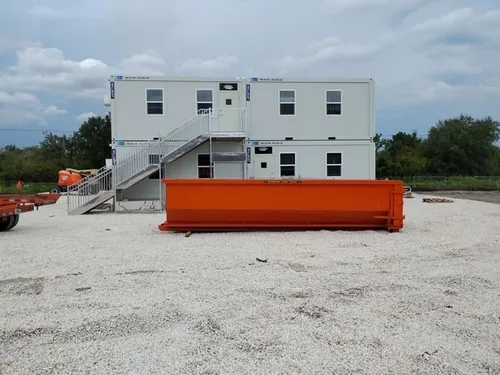 Empty Champs dumpster staged on the gravel lot beside the two story modular office trailers