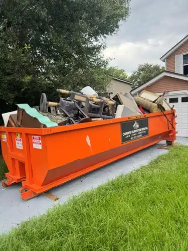 Long Champs dumpster on a Groveland concrete driveway resting on wood boards and loaded with household furniture