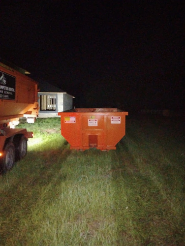 Champs truck lights illuminating a dumpster set on grass beside a block home under construction in Kissimmee at night