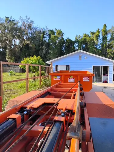 Champs dumpster set on the concrete slab beside the open sliding doors of a light blue Eustis home with a pasture fence and mossy oaks behind