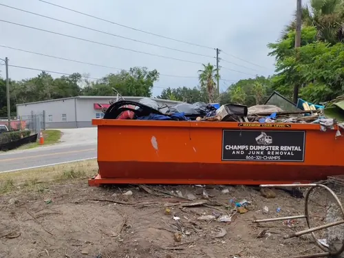 Champs dumpster loaded to the maximum load level line on bare dirt beside a two lane Deltona road with no curb or shoulder
