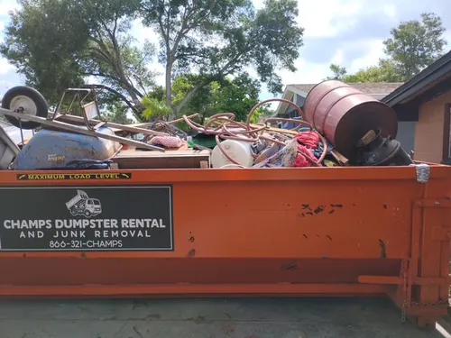Close view of the Champs dumpster loaded to the maximum load level line with spools, hose, a rusted drum, and metal frames